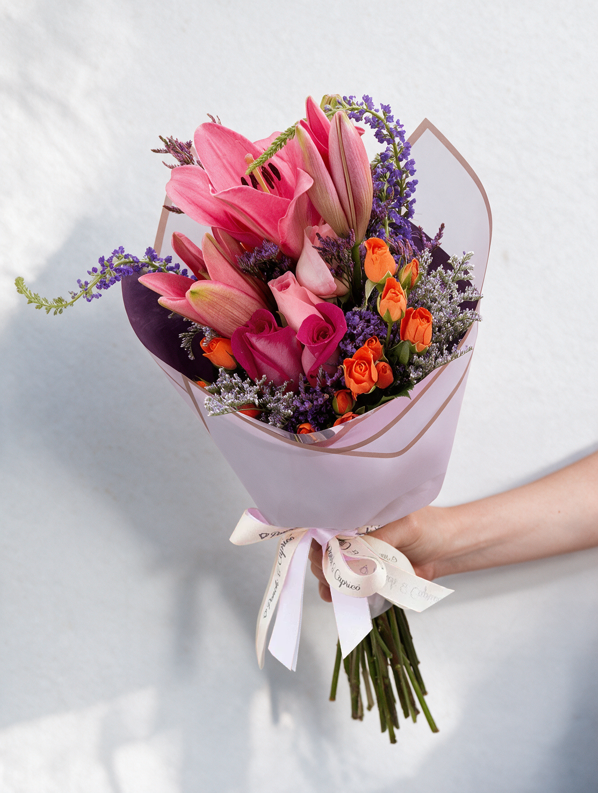 Lindo ramillete atado a mano de Mini Rosas Naranja, Lillies, Rosas Rosada, Rosas Fucsia, Espuma, Verónica y Limonio, envuelto en finos papeles y delicado moño de tela.  Viene incluido florero de vidrio, y caja que cubre el florero (esta última únicamente en la presentación sencilla).