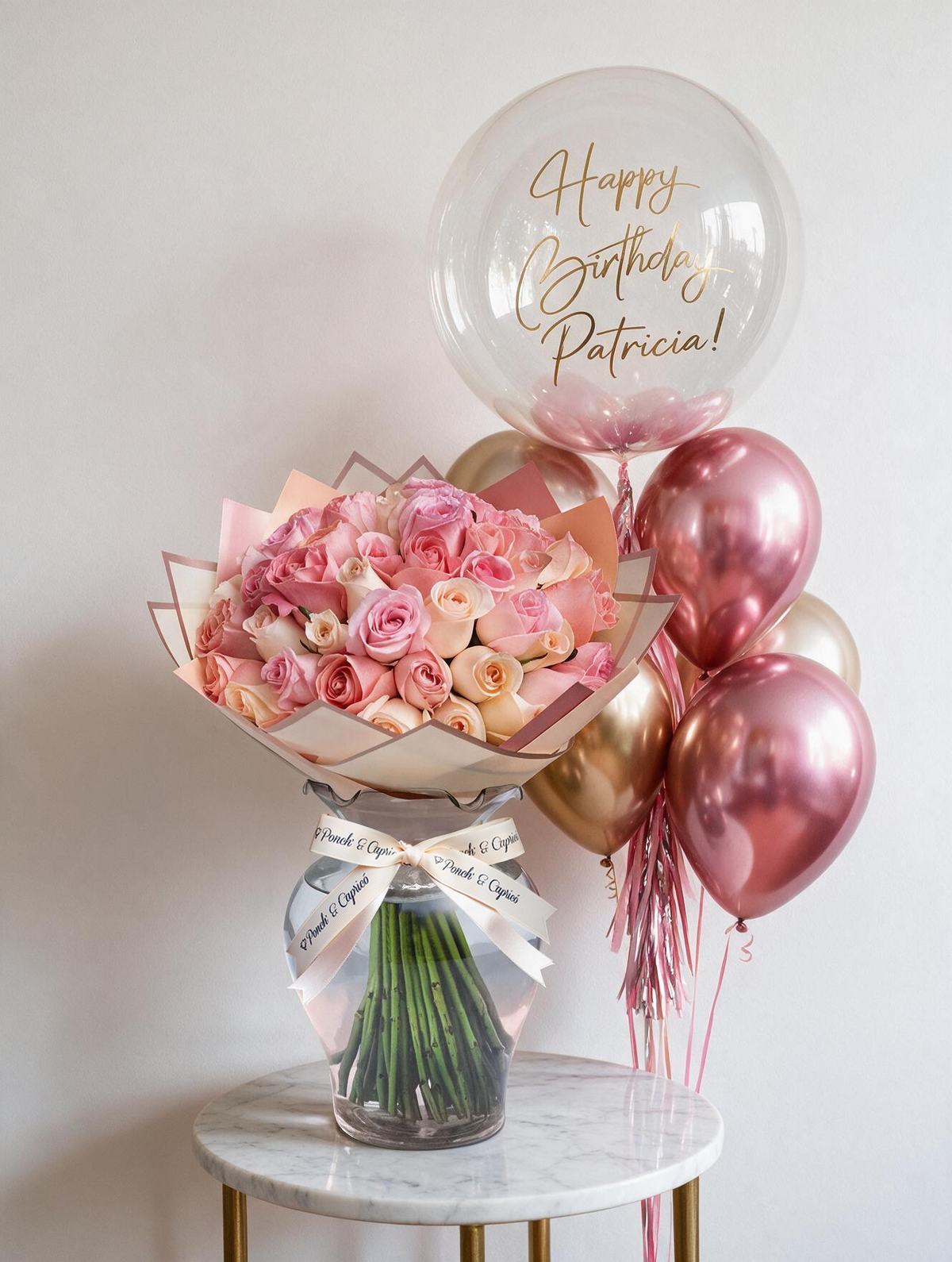 Set de regalo: ramo de 50 rosas rosadas en florero de vidrio con listón Ponch & Capricó junto a una burbuja personalizada con nombre en vinil dorado y globos cromo rosa y oro con helio