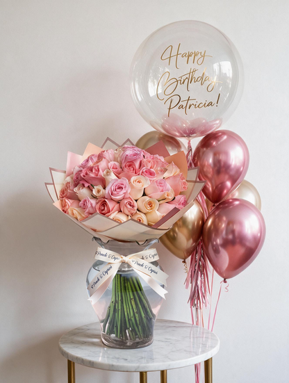 Set de regalo: ramo de 50 rosas rosadas en florero de vidrio con listón Ponch & Capricó junto a una burbuja personalizada con nombre en vinil dorado y globos cromo rosa y oro con helio